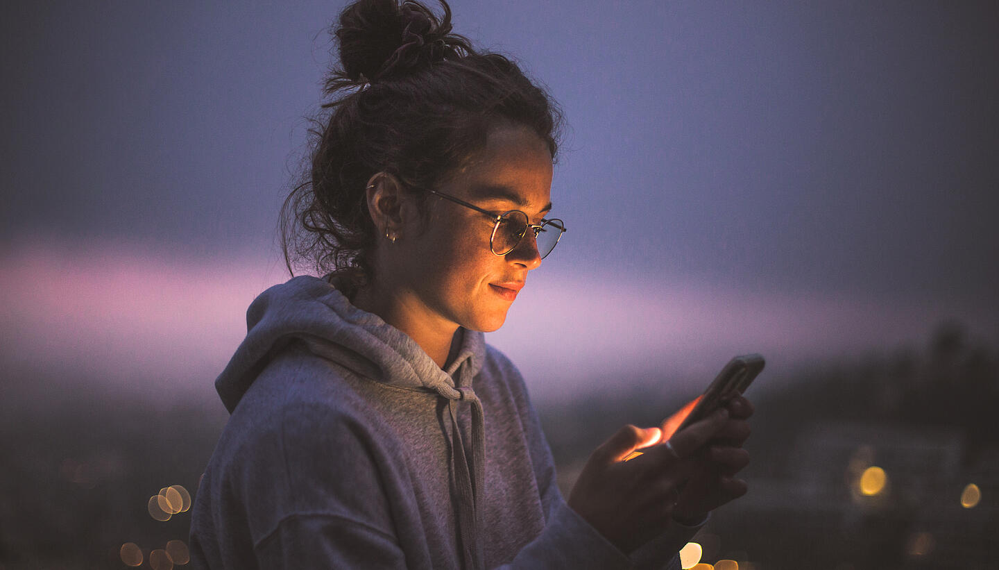 Eine Frau schaut im Dunkeln draußen auf ihr beleuchtetes Smartphone und lächelt. 