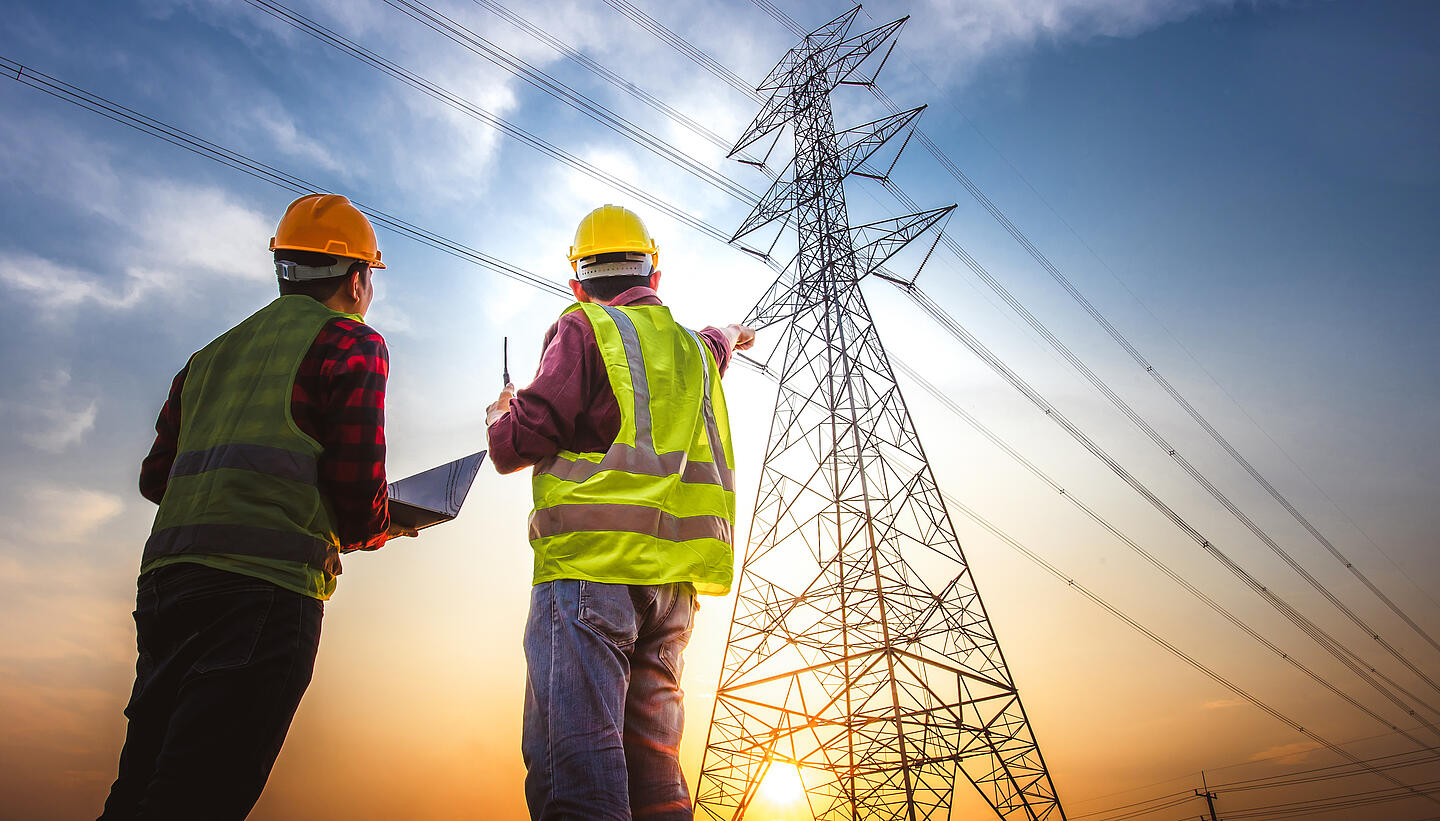 Zwei Männer mit Weste und Helm schauen hoch auf einen Strommast.